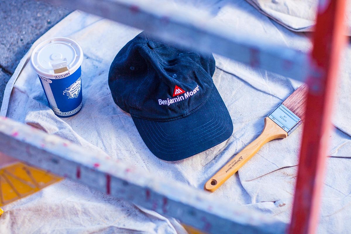 navy blue Benjamin Moore ball cap next to a paint brush and a cup of coffee on a job site. Drop cloth and scaffolding displayed in the background. i